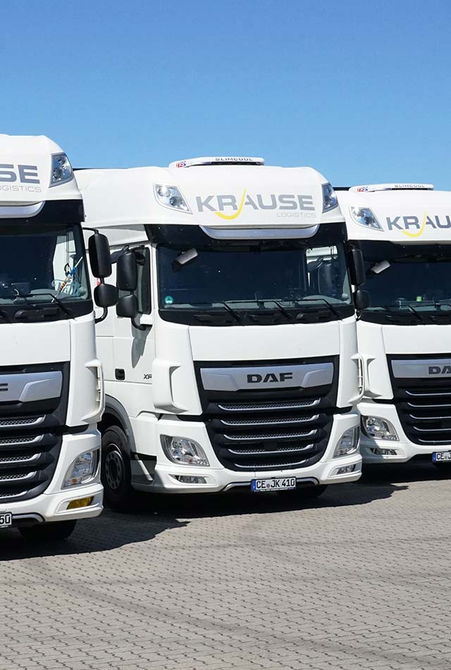 Transportlösungen : A row of white DAF trucks with Krause branding on the wind deflectors, parked on a paved surface under a clear blue sky.